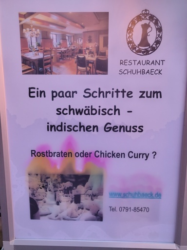 Ein Plakat, das ein Titelbild eines Restaurantsinnenraums zeigt. Daneben ist ein stilisierter Bierkrug eingefasst in Ornamenten, dass das Logo des Restaurants Schuhbaeck zeigt. Dem folgt der Text: "Ein paar Schritte zum schwäbisch - indischen Genuss Rostbraten oder Chicken Curry ?" Abschließend ist ein weiteres Bild eines Gedecks und Kontaktdaten ausgeführt. Der Druck ist von Feuchtigkeit verwaschen.