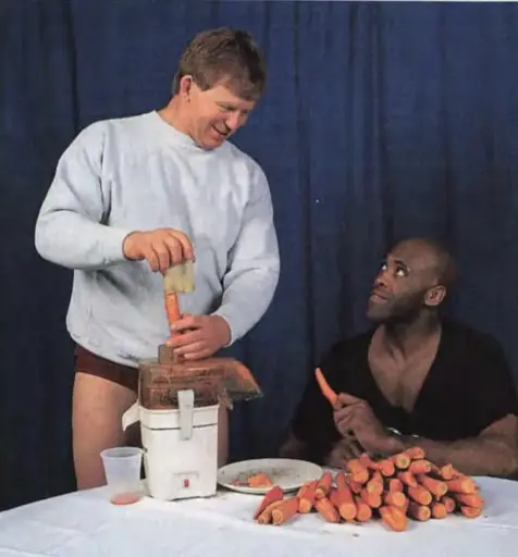 Bob Backlund stands behind a table in a white sweatshirt and speedo, he's cramming a carrot into a juicier. He smiles at Virgil who is seated next to him. Virgil looks up at Bob with a weird smirk. There are about 40 thousand carrots on the table in front of them. I dunno it's like 30 or something. It's way too many carrots!