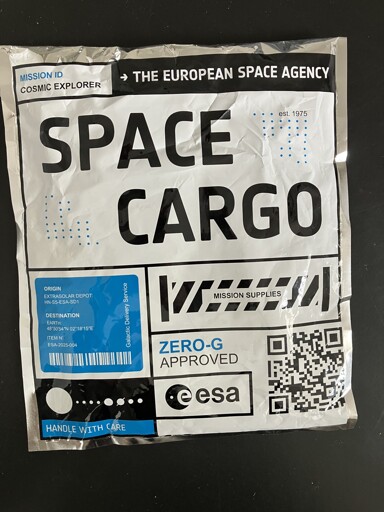A silver wrapped parcel with the words "SPACE CARGO" in bold letters. Elsewhere are decals saying :


Mission ID  cosmic explorer
-> The European space agency
| mission supplies |
[origin extra solar depot HN-S5-ESA-SD1
Destination Earth
48 50 54N 02 18 15E
Item no ESA-2025-004
Galactic delivery service]

HANDLE WITH CARE

Zero-G Approved

ESA

