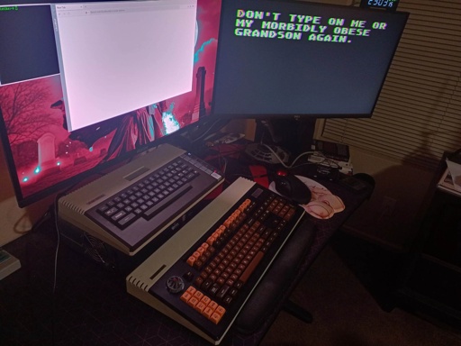 A view of a desk with two monitors.  A vintage Atari 800XL computer is in the back, driving one monitor displaying the text "Don't type on me or my morbidly obese grandson again."  in front is a custom 130% PC keyboard with similar design language and colours.