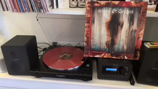 Picture of an LP sleeve balanced on a hi-fi system, with the vinyl record playing on a turntable next to the hi-fi system