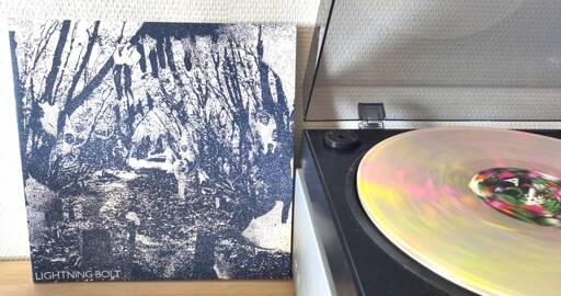 The album cover (left) is a high contrast monochrome, abstract forest setting(?) printed in a desaturated cobalt blue on white. The vinyl spinning on the turntable (right) is much more colourful with almost fluorescent yellow and pink splatter melted into the clear vinyl.