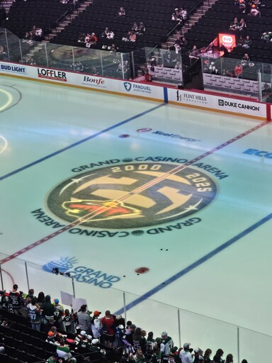 A high-angle view of an ice hockey rink with a large 'Grand Casino Arena 2000-2025' anniversary logo at center ice, incorporating the Minnesota Wild logo, as seen from the spectator stands
